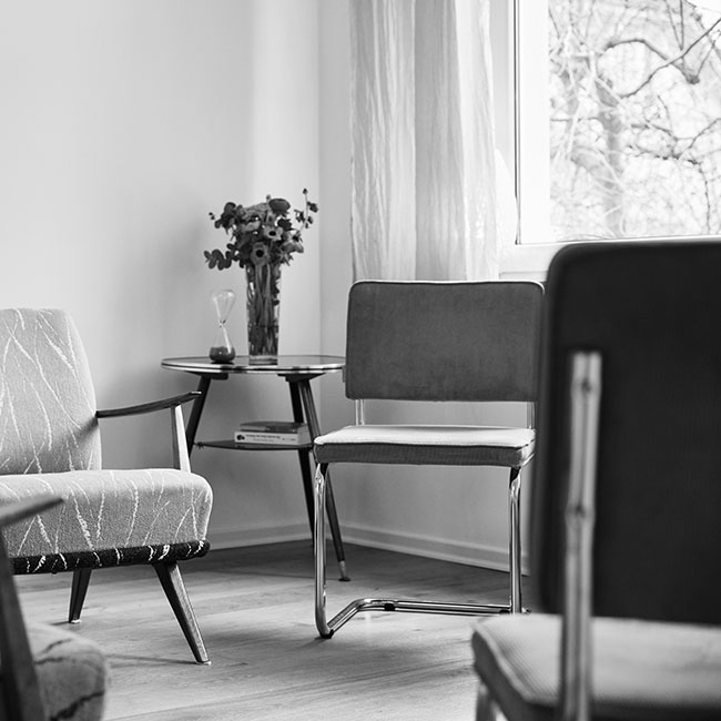 Raumausschnitt mit Stühlen, die in einem Kreis stehen, in der Ecke dahinter ein kleiner Tisch mit einem Blumenstrauß, seitlich ein Fenster mit hellem Vorhang