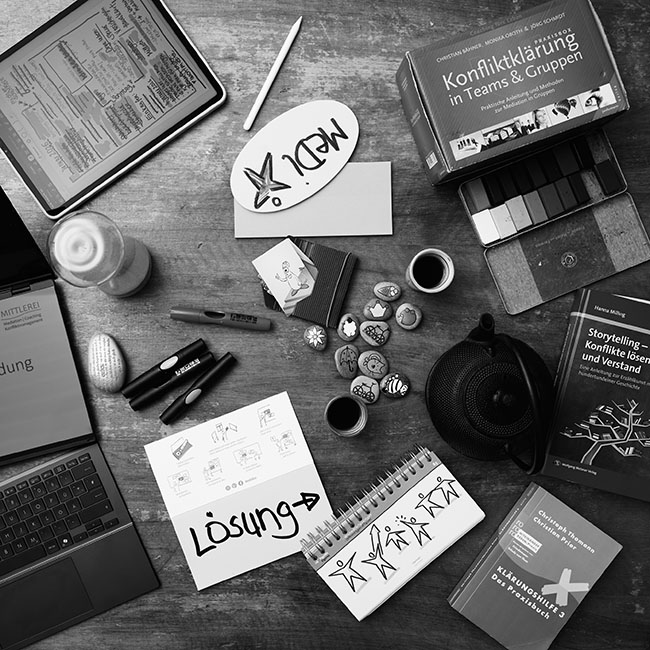 Tisch mit vielen Arbeitsmaterialien zum Thema Mediation von oben gesehen: Stifte, Papier, Karten, Wachsmalblöcke, Bücher, Laptop