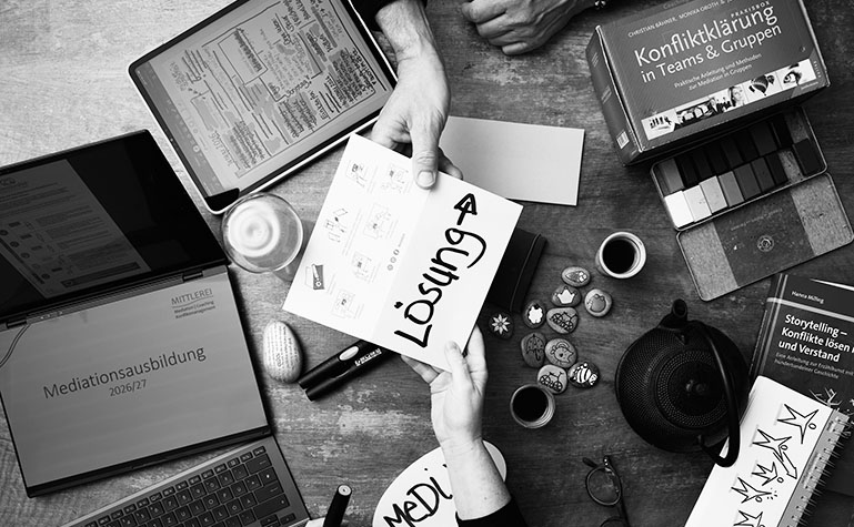 Tisch mit vielen Arbeitsmaterialien zum Thema Mediation von oben gesehen: Stifte, Papier, Karten, Wachsmalblöcke, Bücher, Laptop, von unten und oben zeigt jeweils eine Hand herein, beide halten zusammen eine Karte mit der Aufschrift Lösung