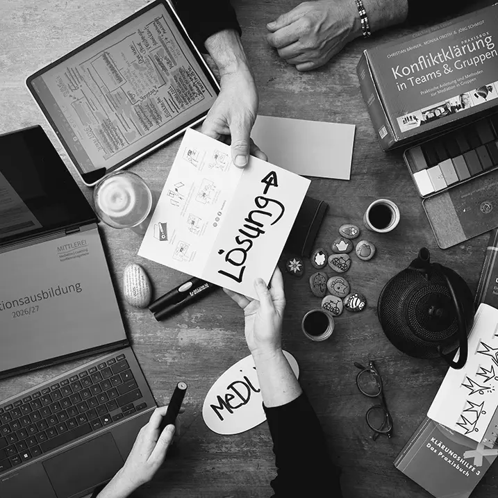 Tisch mit vielen Arbeitsmaterialien zum Thema Mediation von oben gesehen: Stifte, Papier, Karten, Wachsmalblöcke, Bücher, Laptop, von unten und oben zeigt jeweils eine Hand herein, beide halten zusammen eine Karte mit der Aufschrift Lösung